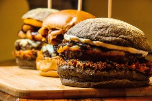 Tre burger gourmet impilati con panino scuro, formaggio filante e bacon, su tagliere di legno